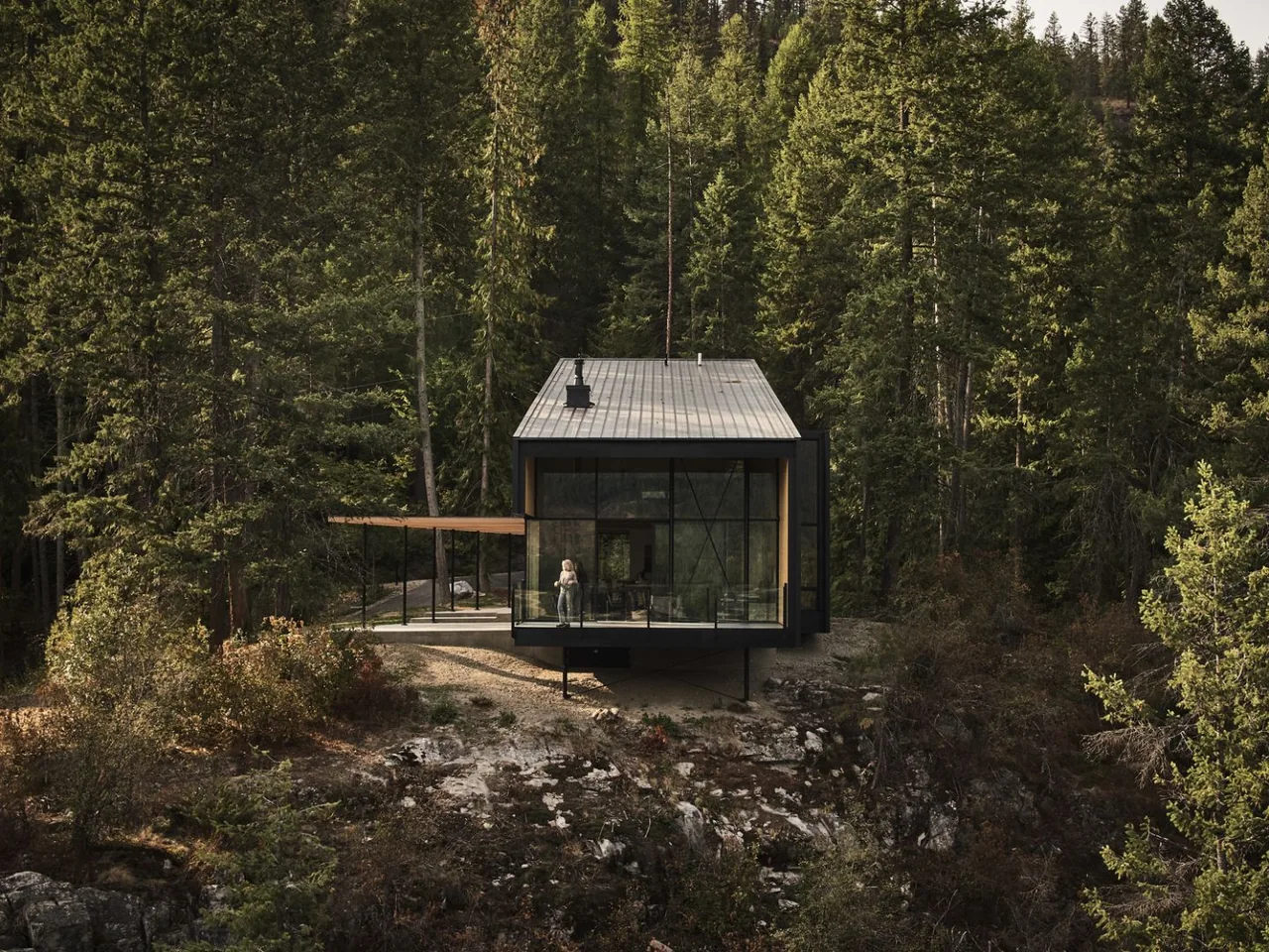 Modern cabin perched on rocky terrain in dense forest with a person standing inside near large glass windows.