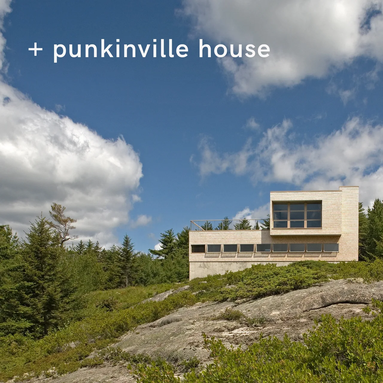 Modern house with white wood siding and concrete base on rocky hillside under blue sky with clouds.
