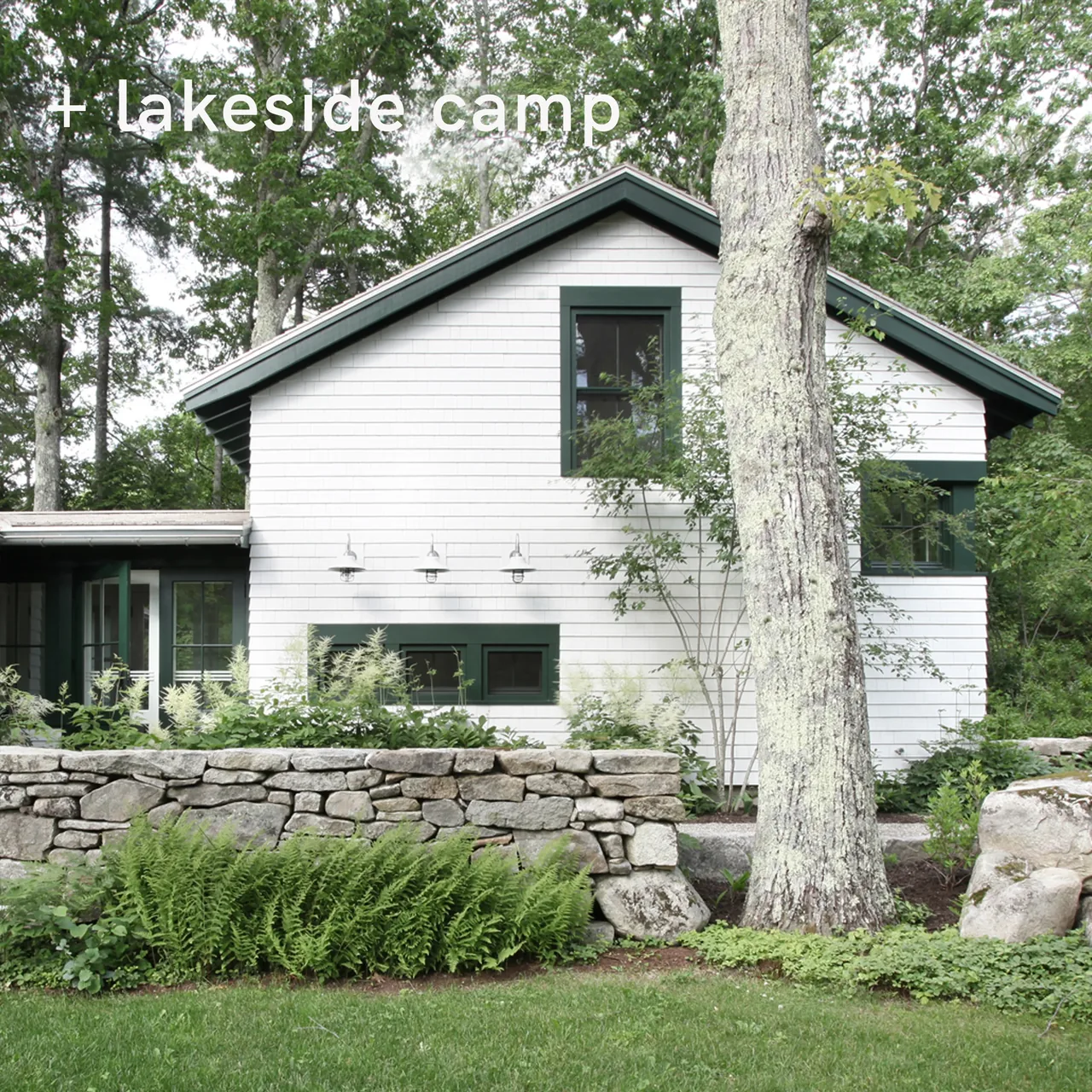 White house exterior with shingle siding, green-framed windows, stone wall, and surrounding greenery.