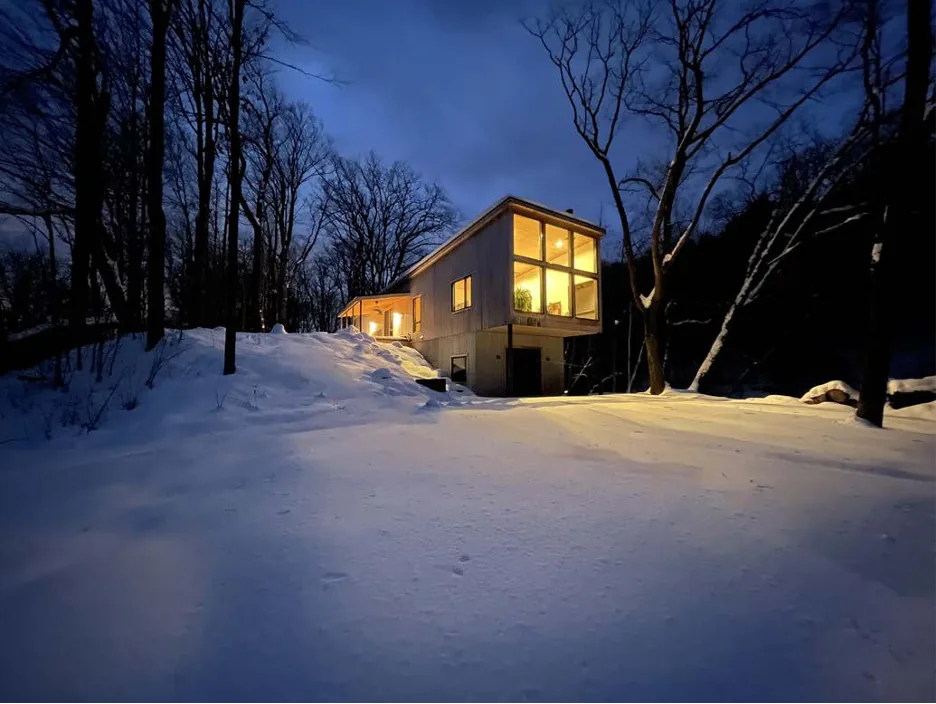 A modern house glowing from interior lights set in a snowy wooded area at dusk.