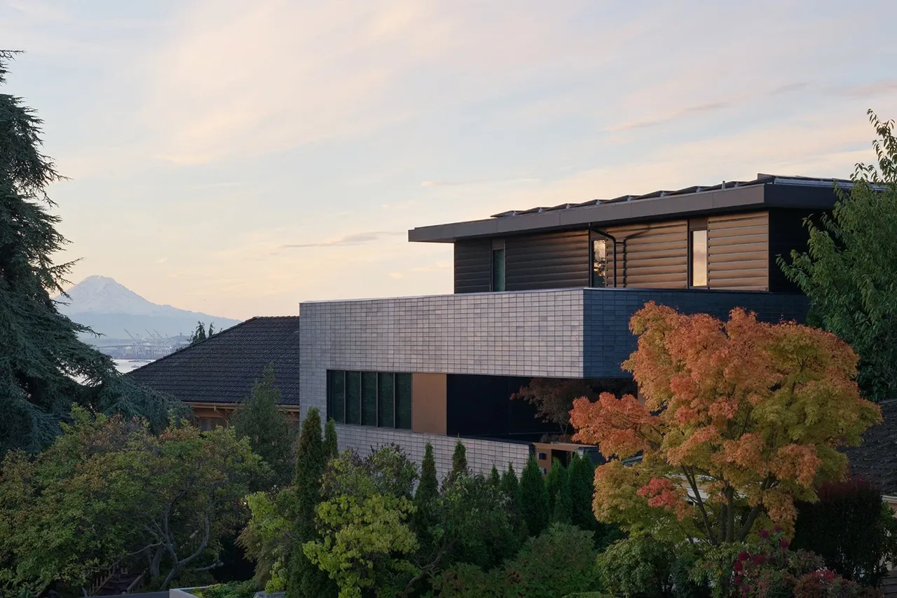 Modern two-story house with light-colored brick lower level and dark wood siding upper level, surrounded by trees with mountain in the far,