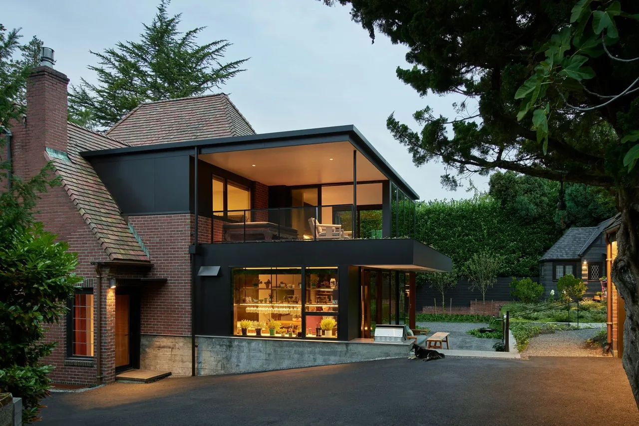 Exterior view of a two-story house combining traditional brick and modern black metal elements with large windows and a balcony at dusk.