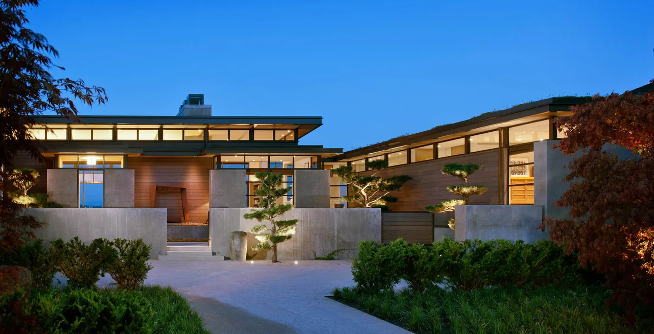 Evening view of a modern residence with illuminated clerestory windows and concrete walls surrounded by trees and shrubs.