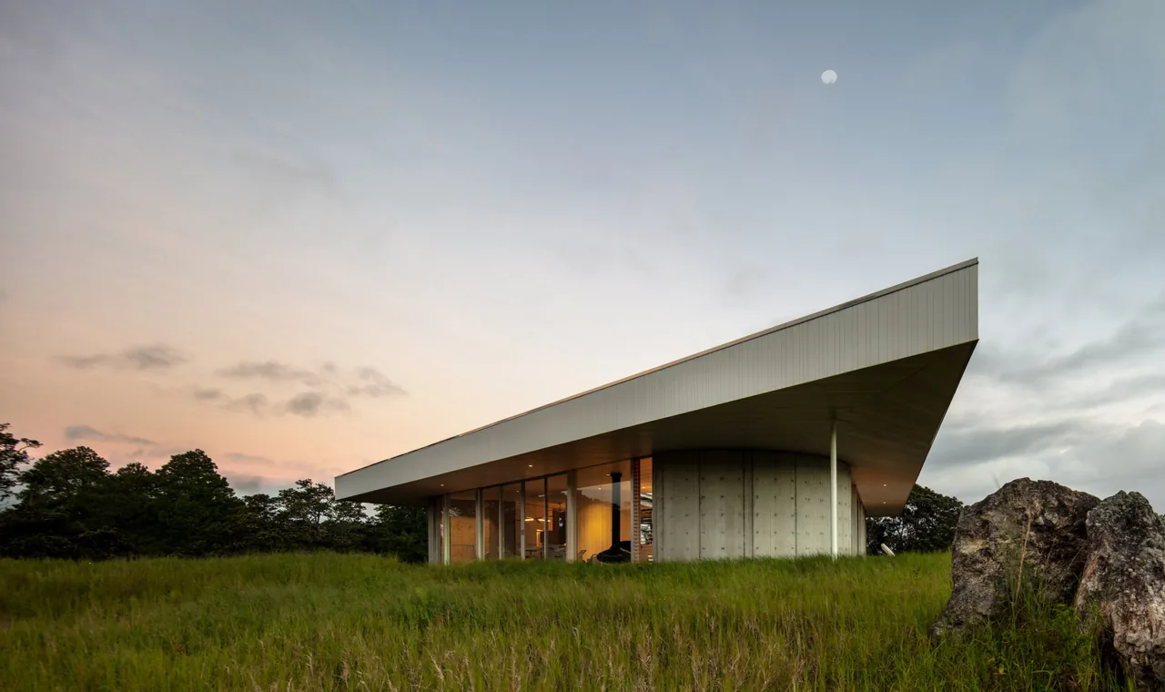 Modern house with sharp angled flat roof and glass walls set in grassy landscape at dusk.