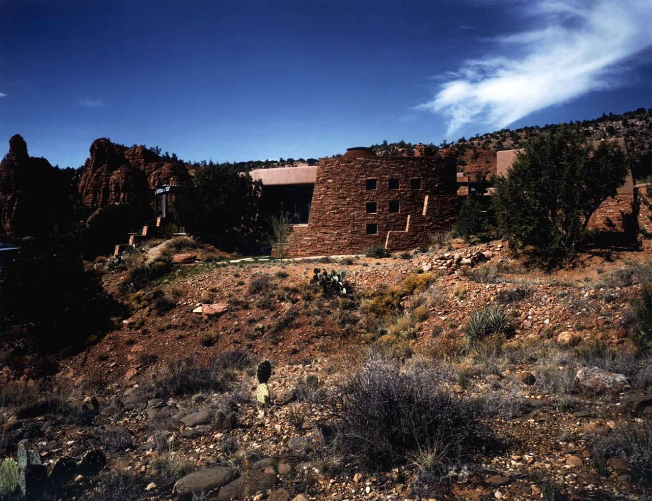 Stone house with small square windows set in a desert landscape with rocky terrain and sparse vegetation under a blue sky.