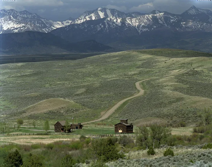 Two wooden farm buildings on a green ranch landscape with a winding dirt road and snow-capped mountains in the background.