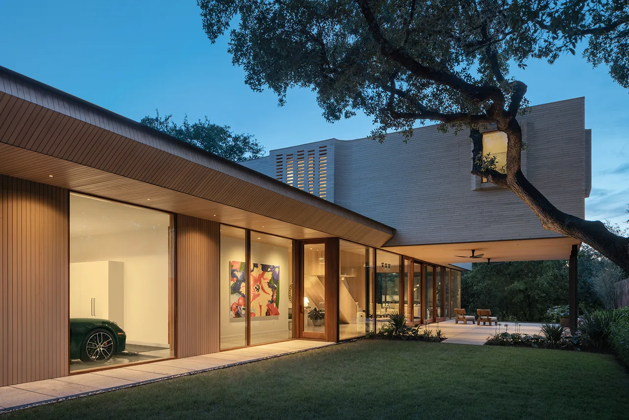 Modern two-story house exterior in evening with large glass windows and overhanging upper floor near a large tree.