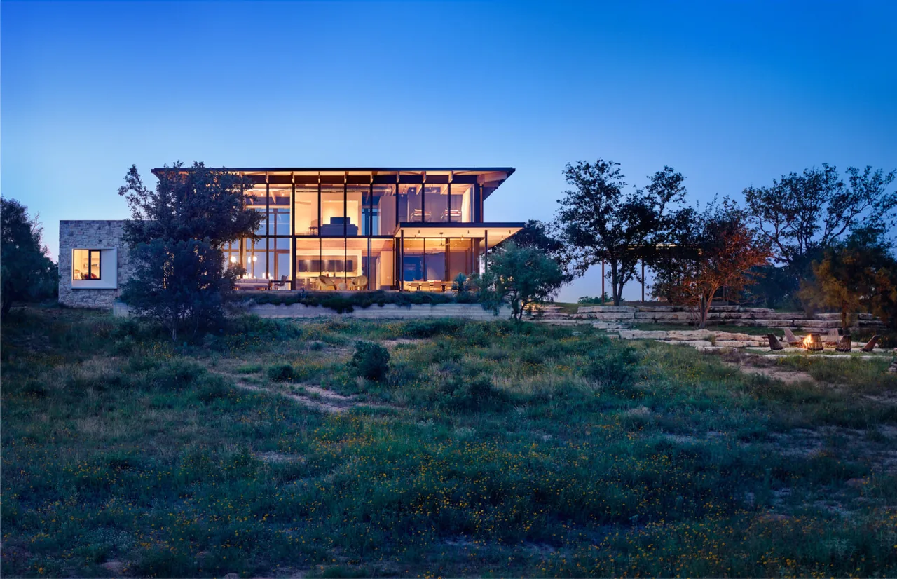 Modern two-story house with extensive floor-to-ceiling glass walls, illuminated at dusk and surrounded by trees and grassy landscape.
