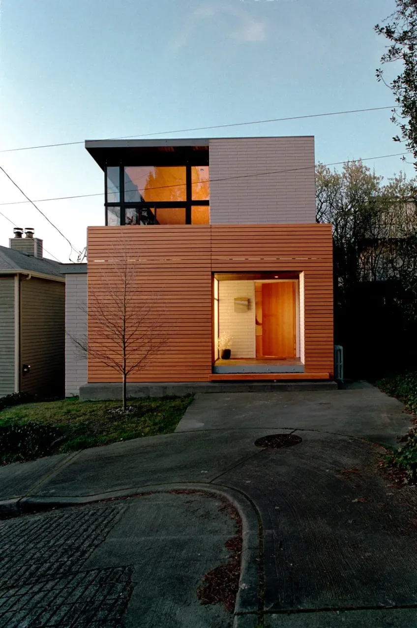 Modern two-story house with wood siding and large upper floor windows facing the street at dusk.
