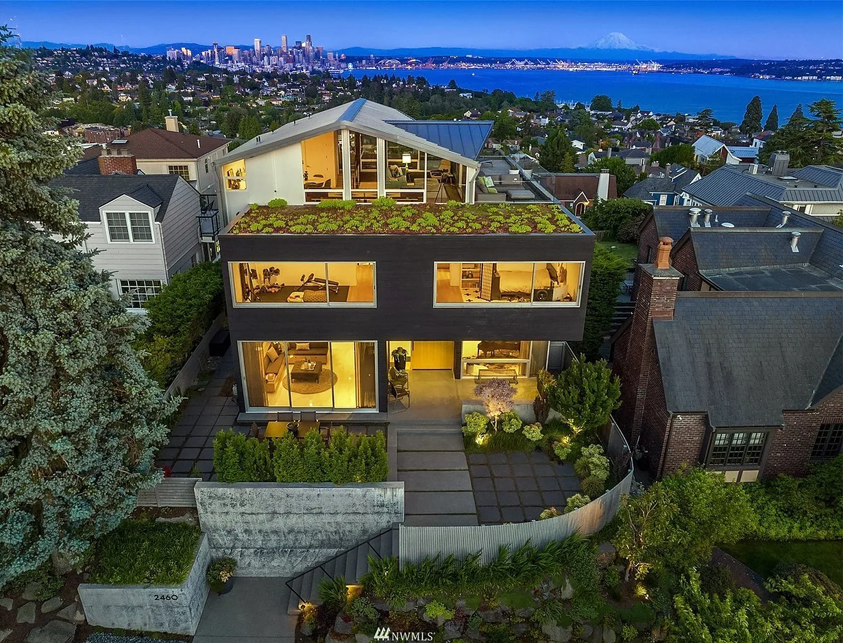 Modern three-story house at dusk with large windows, a green roof, and city skyline in the background.