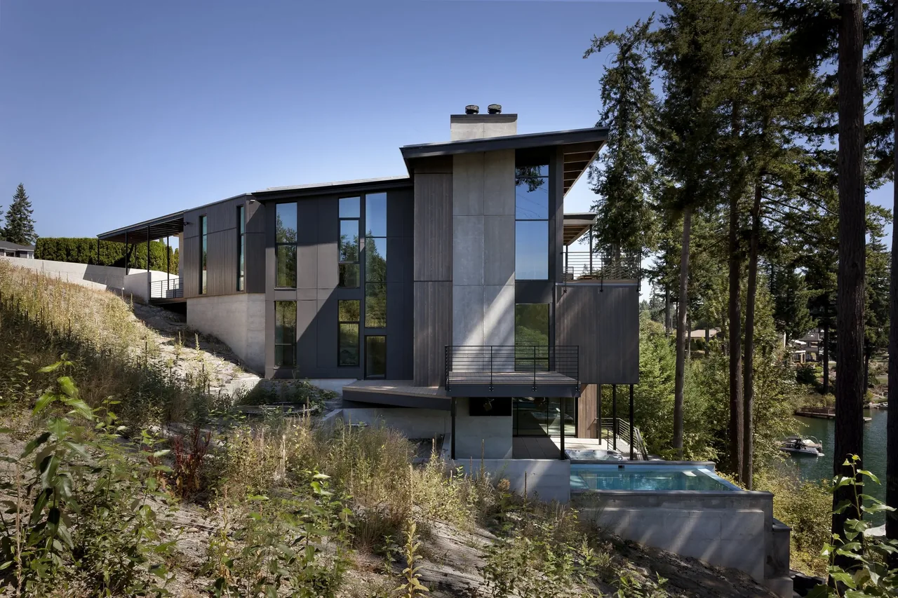 Modern multi-level house with large vertical windows overlooking a lakeside forested area and an infinity pool.