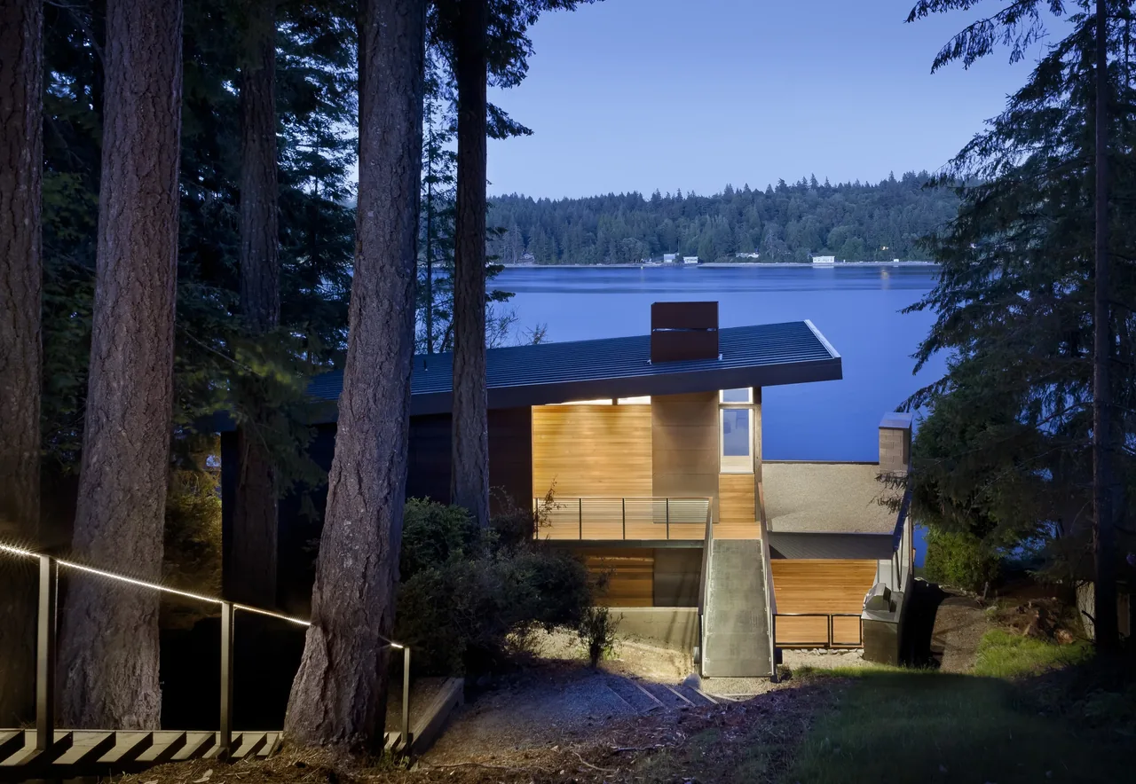 Evening view of a modern cabin with a sloped roof beside a large body of water, seen through tall trees.