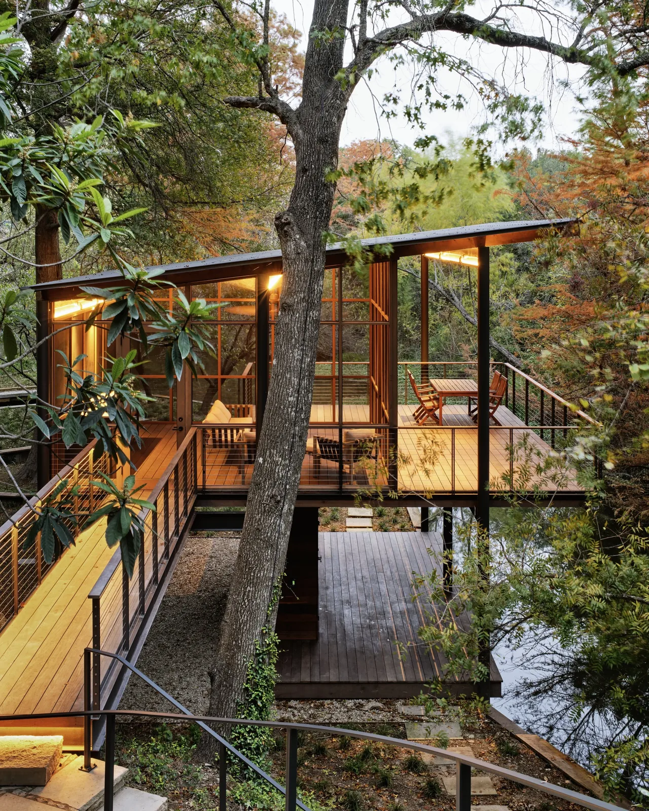 A modern elevated house with floor-to-ceiling glass walls surrounded by trees beside a water body in autumn.