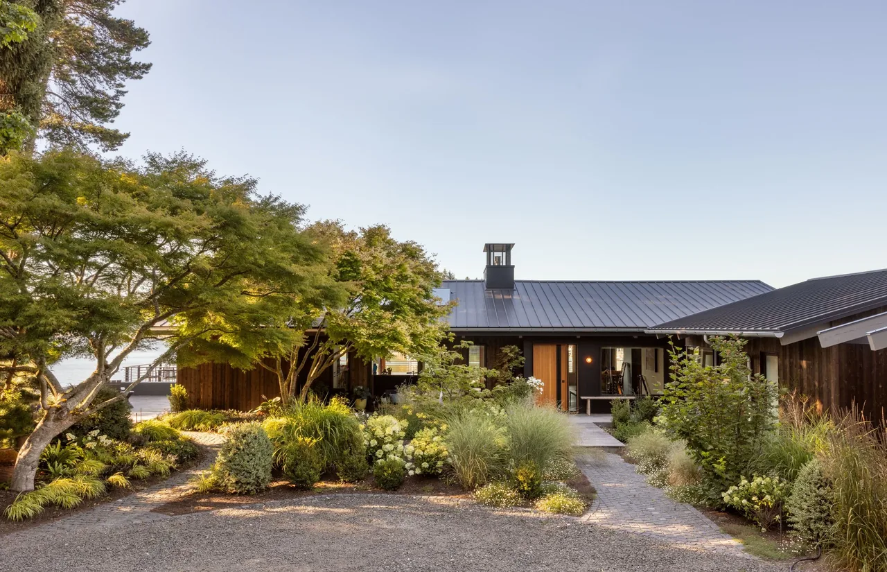 Front view of a modern house with a metal standing seam roof surrounded by lush landscaping and trees under a clear sky.