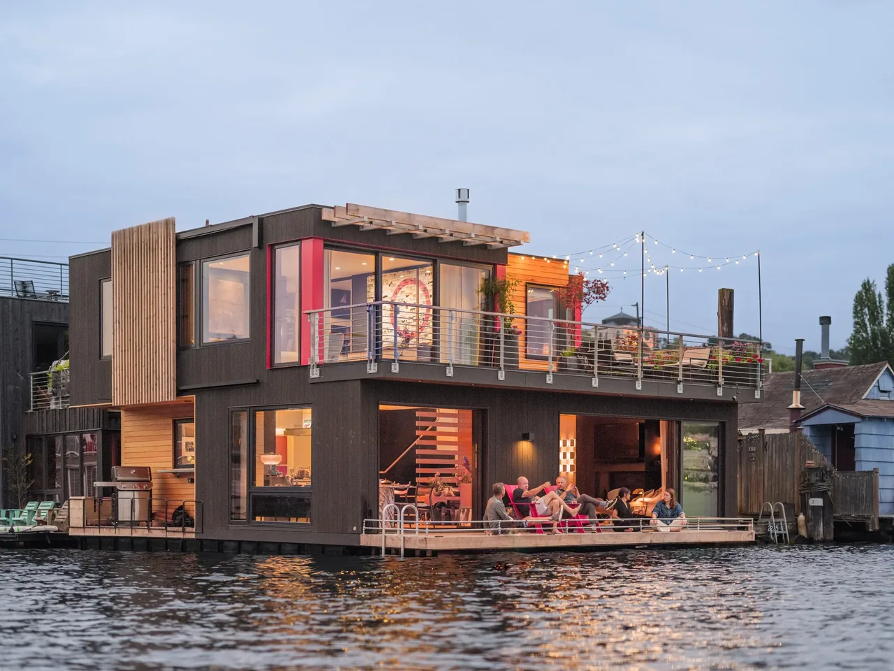 Modern two-story floating home with dark siding, wooden accents, and open deck by the water at dusk.