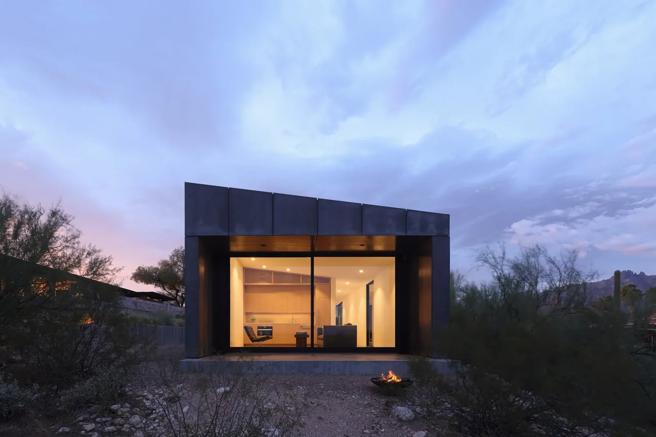 Modern small house with large floor-to-ceiling window showing interior living space at dusk in desert landscape.