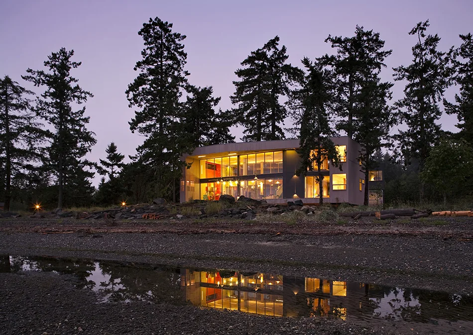 Modern beach house illuminated in evening surrounded by tall trees and reflected in water on rocky shore.