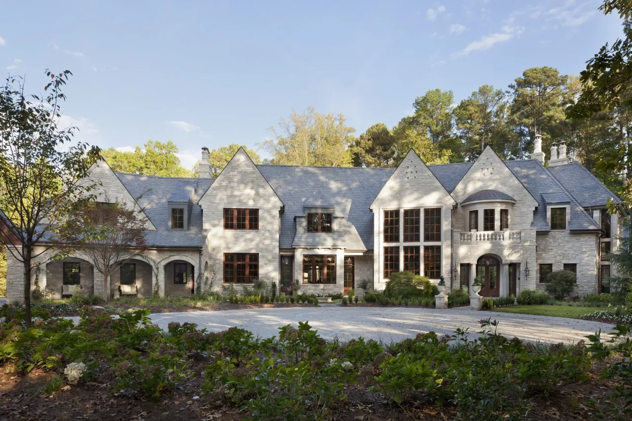Front view of a large stone house with steep gable roofs and arched entryways in a wooded setting.