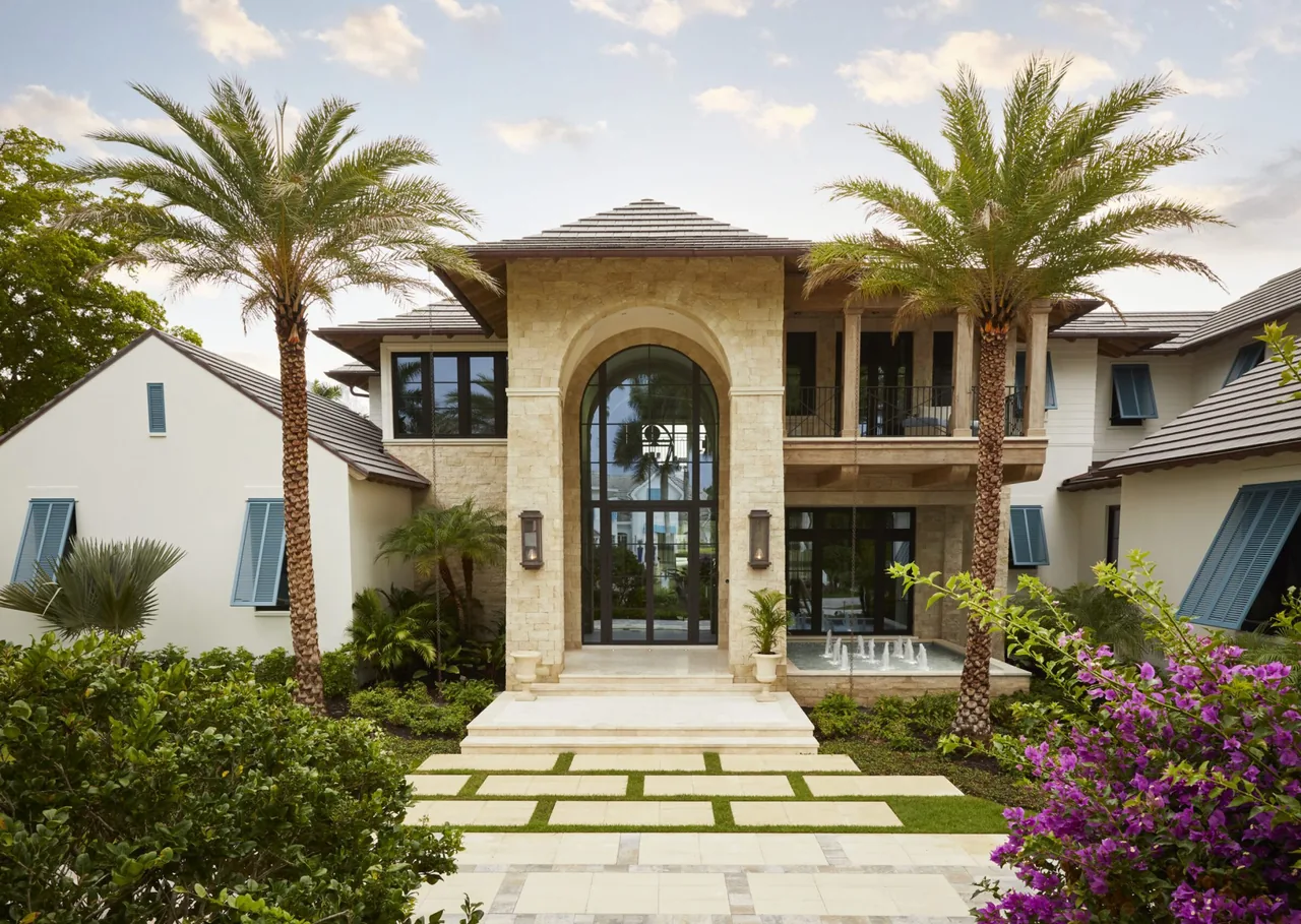 Front exterior of a two-story coastal transitional house with a large arched glass front door, stone and white siding, flanked by palm trees