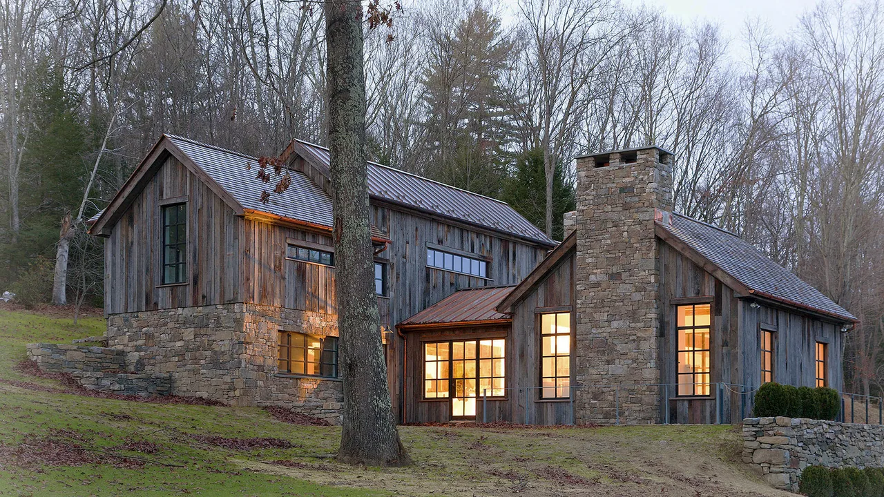 Exterior view of a two-story house with stone and wood siding, illuminated windows, and a tall stone chimney at dusk.