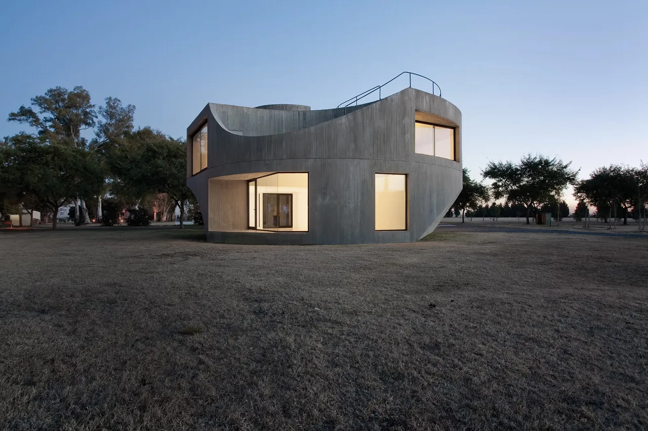 Curved concrete house exterior at dusk with large windows and minimal landscaping.