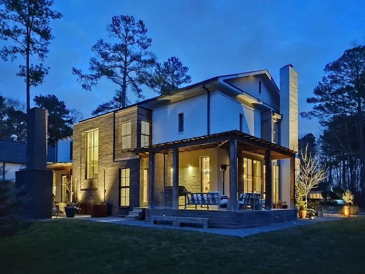 Evening view of a modern two-story residence with illuminated windows and a covered patio surrounded by tall trees.