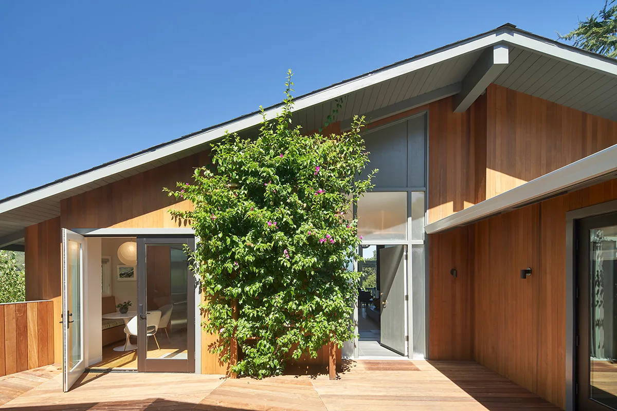 Exterior view of a modern wood-paneled house with a slanted roof and an open double door leading to an interior room, featuring a lush green