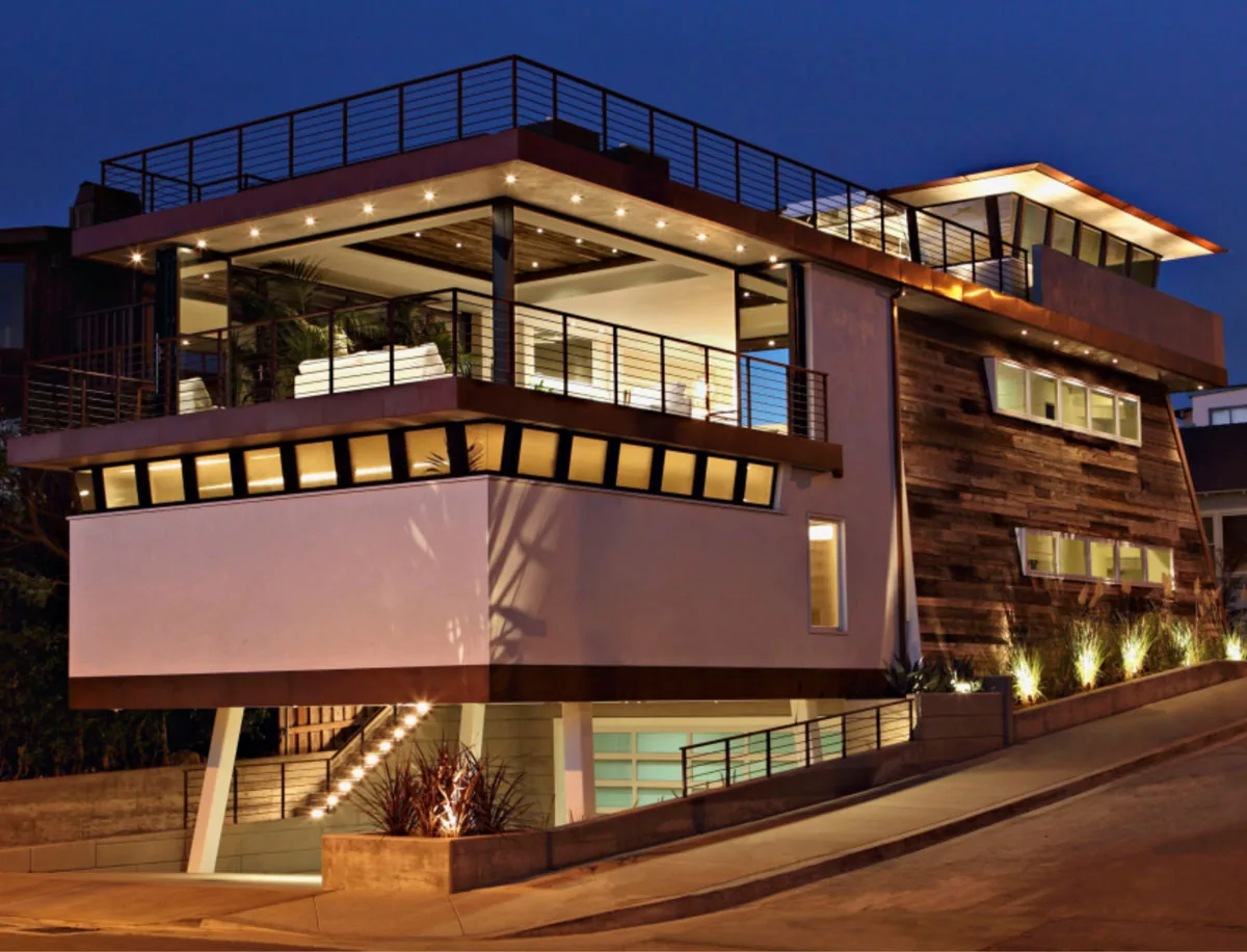 Modern multi-story house with large windows and outdoor balcony, lit up at dusk on a sloped street.