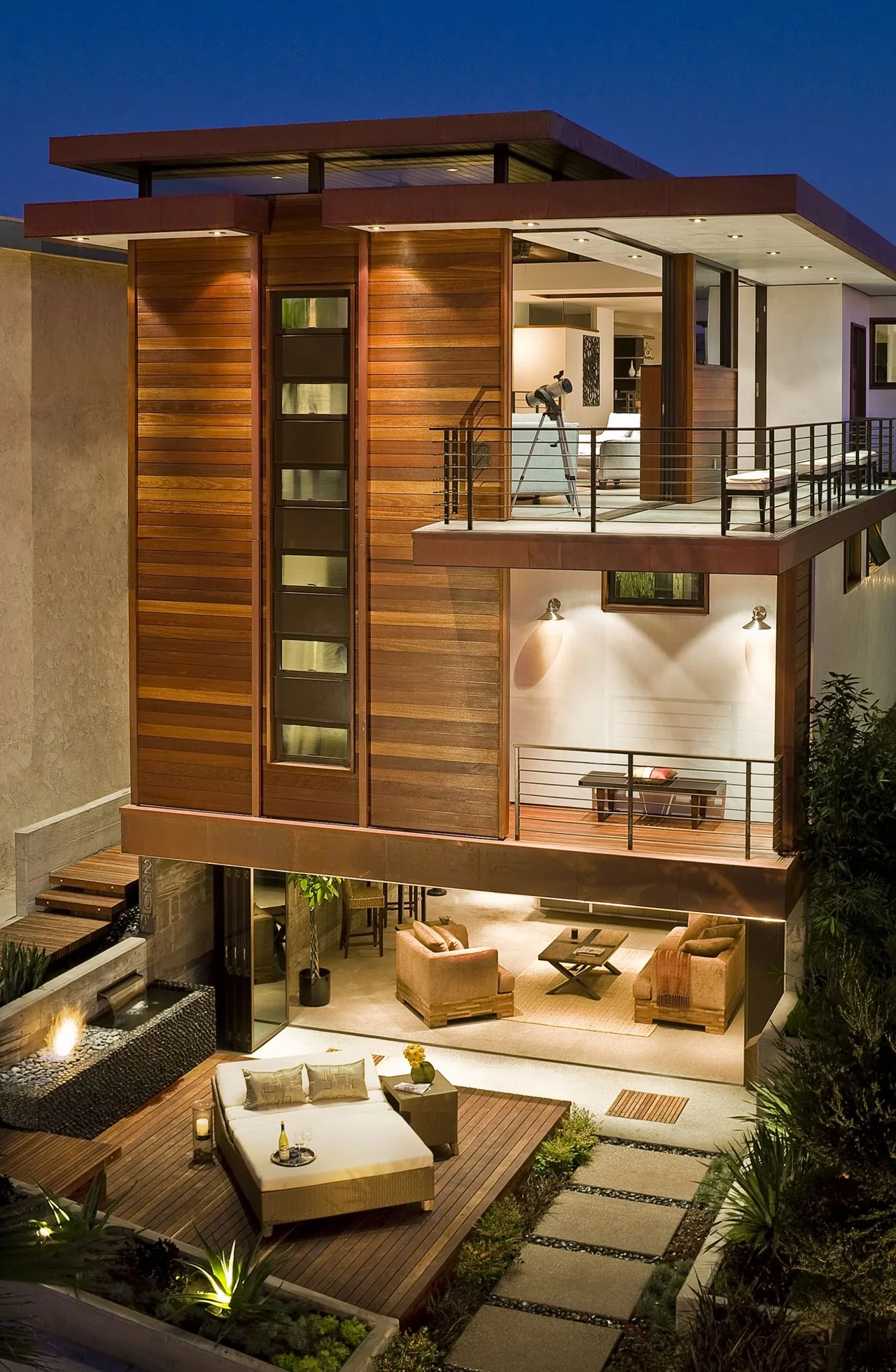 Evening view of a modern two-story home with wood siding, metal railings, and outdoor seating areas with lighting.