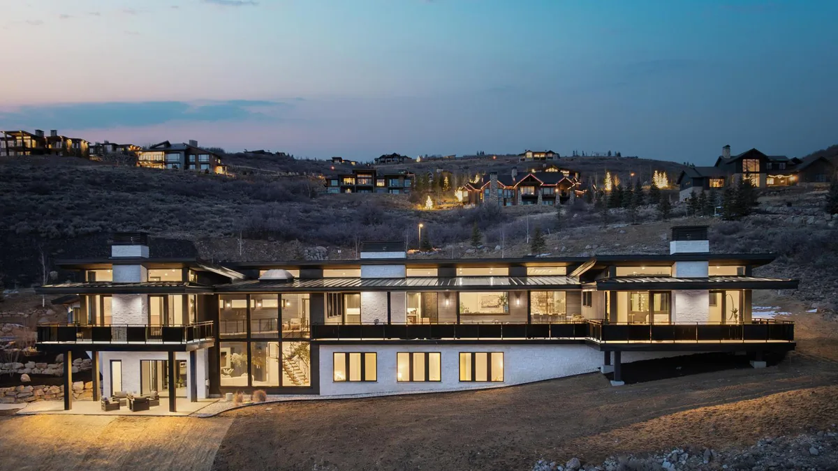 Modern house exterior at dusk with lights on, situated on a hillside with multiple large windows and balconies.