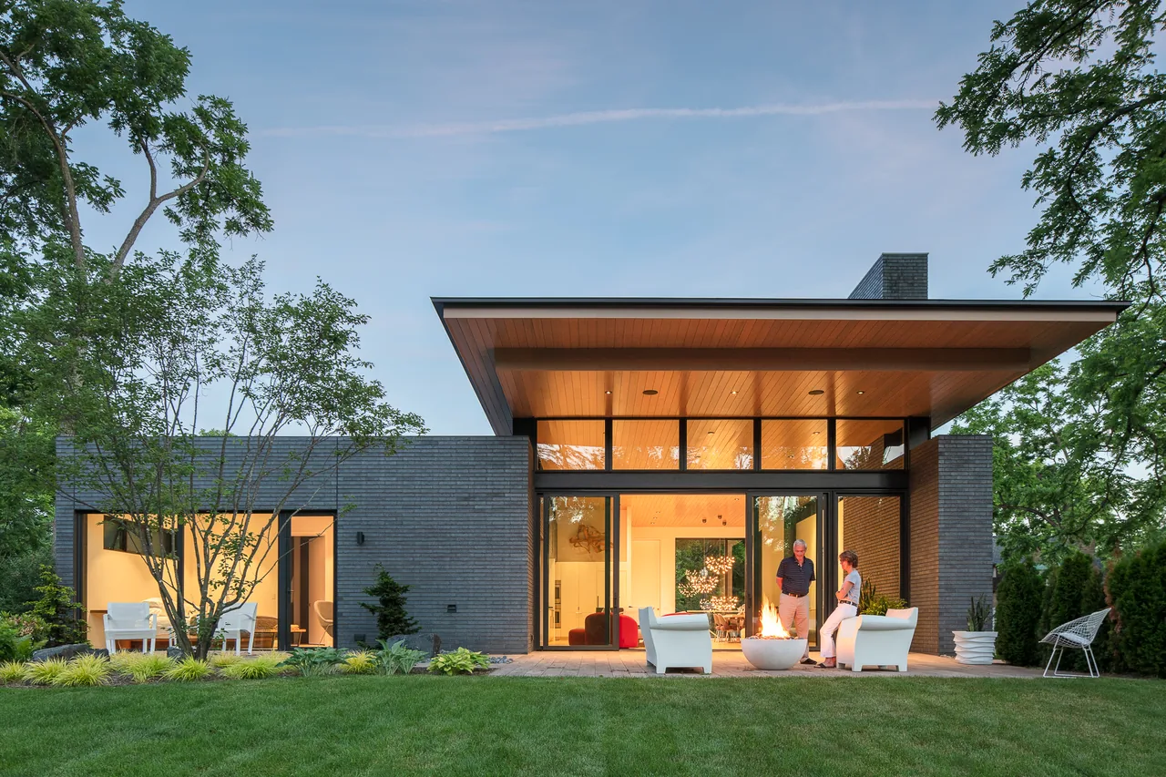 Modern single-story house exterior with dark brick walls, large floor-to-ceiling windows, and a wooden overhang at twilight.