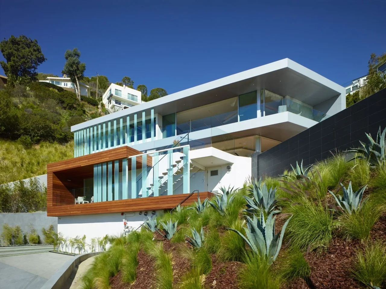 Modern multi-level house on a hillside with white and wood exterior and glass railings under a clear blue sky.