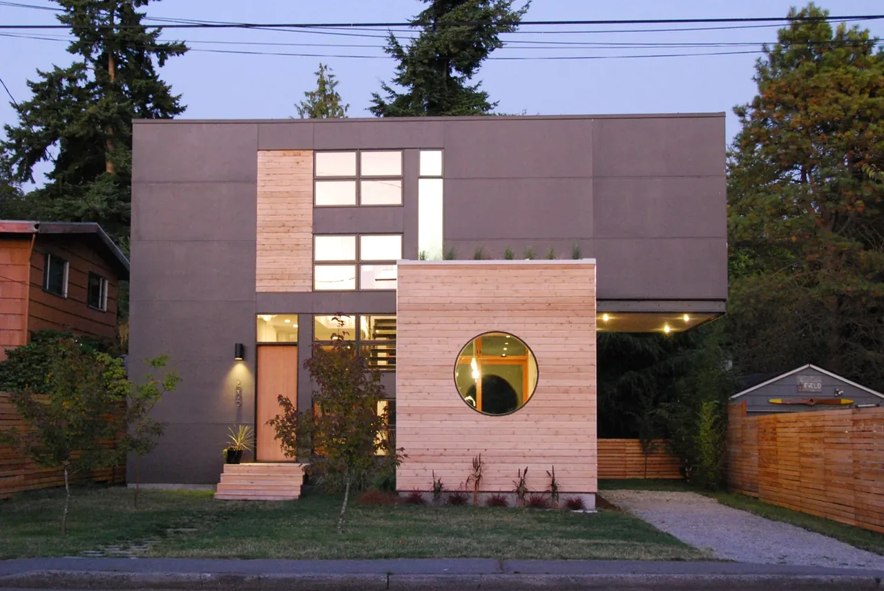 Front exterior of a modern two-story house with mixed wood and dark panel siding and a round window on the wooden section.