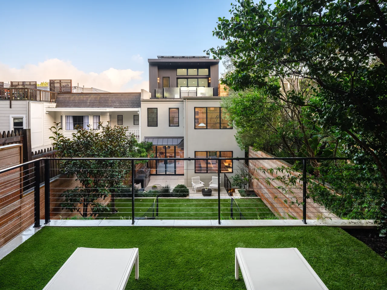 View of a modern multi-story residence from an upper balcony with green turf and two white lounge chairs.