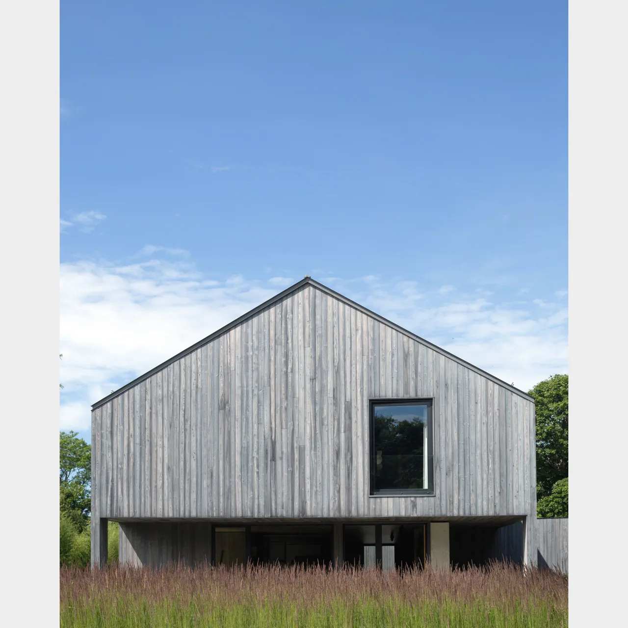 Exterior view of a modern gable-roof house with vertical wood siding and a single window above an open ground level.