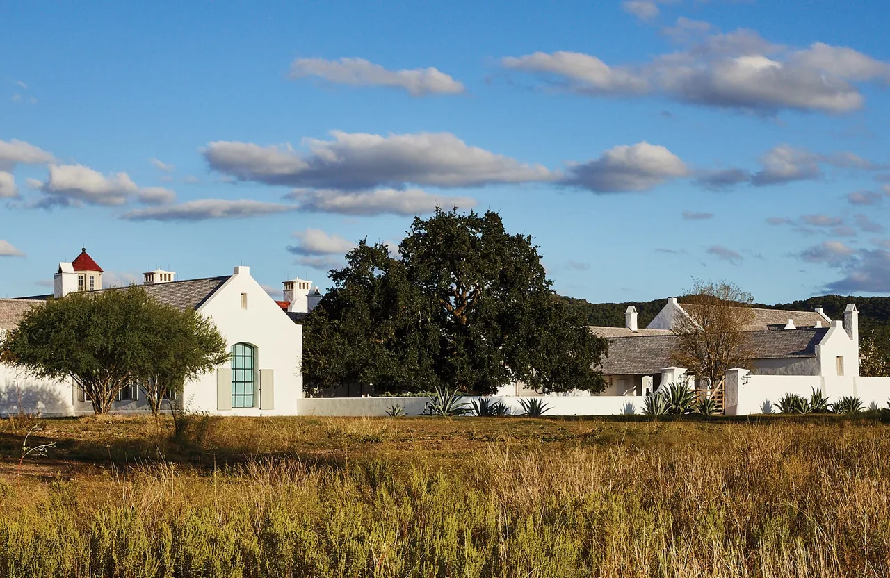 Wide exterior view of a white farmhouse-style home with pitched roofs and multiple chimneys under a partly cloudy blue sky.