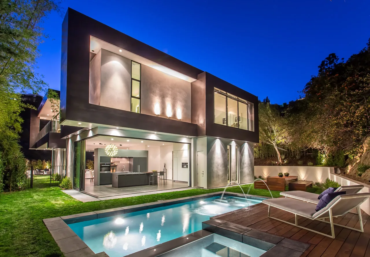 Night view of a modern two-story home with an open kitchen, pool, and outdoor lounge seating on a wooden deck.