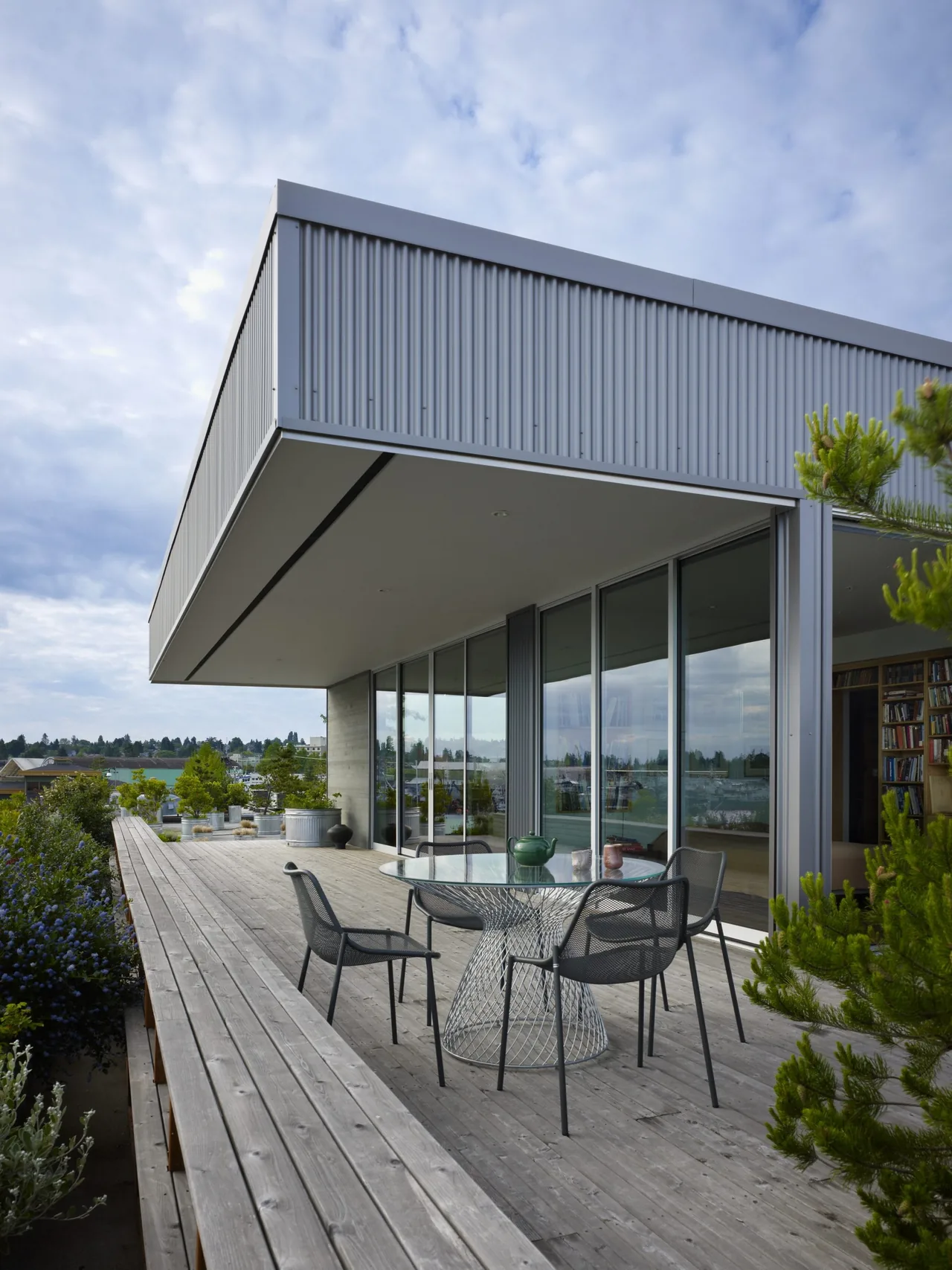 Outdoor deck with metal dining table and chairs beside a modern home with large glass sliding doors under an overhanging roof.