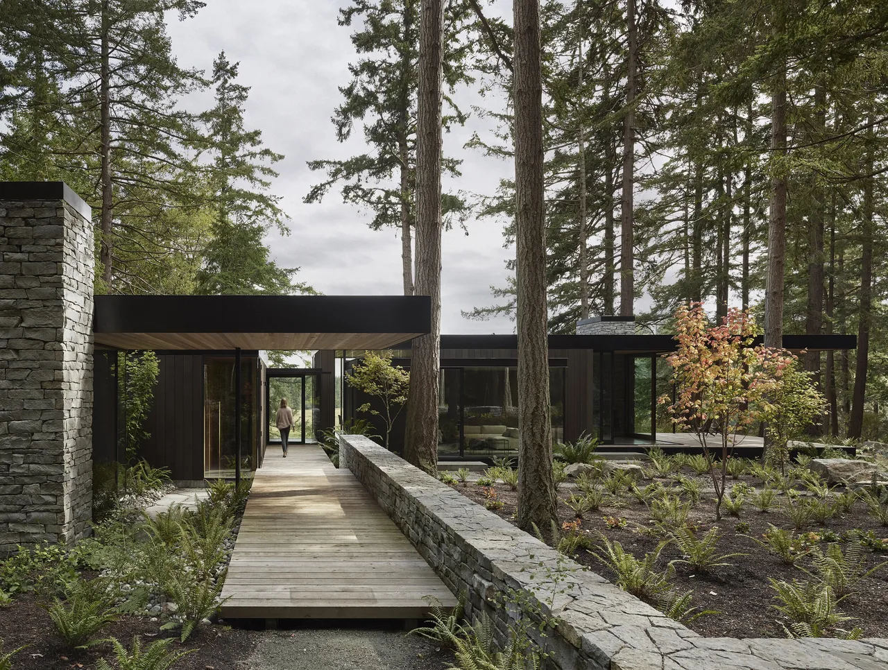 Modern single-story house in a wooded setting with stone wall and wood walkway leading to the entrance.