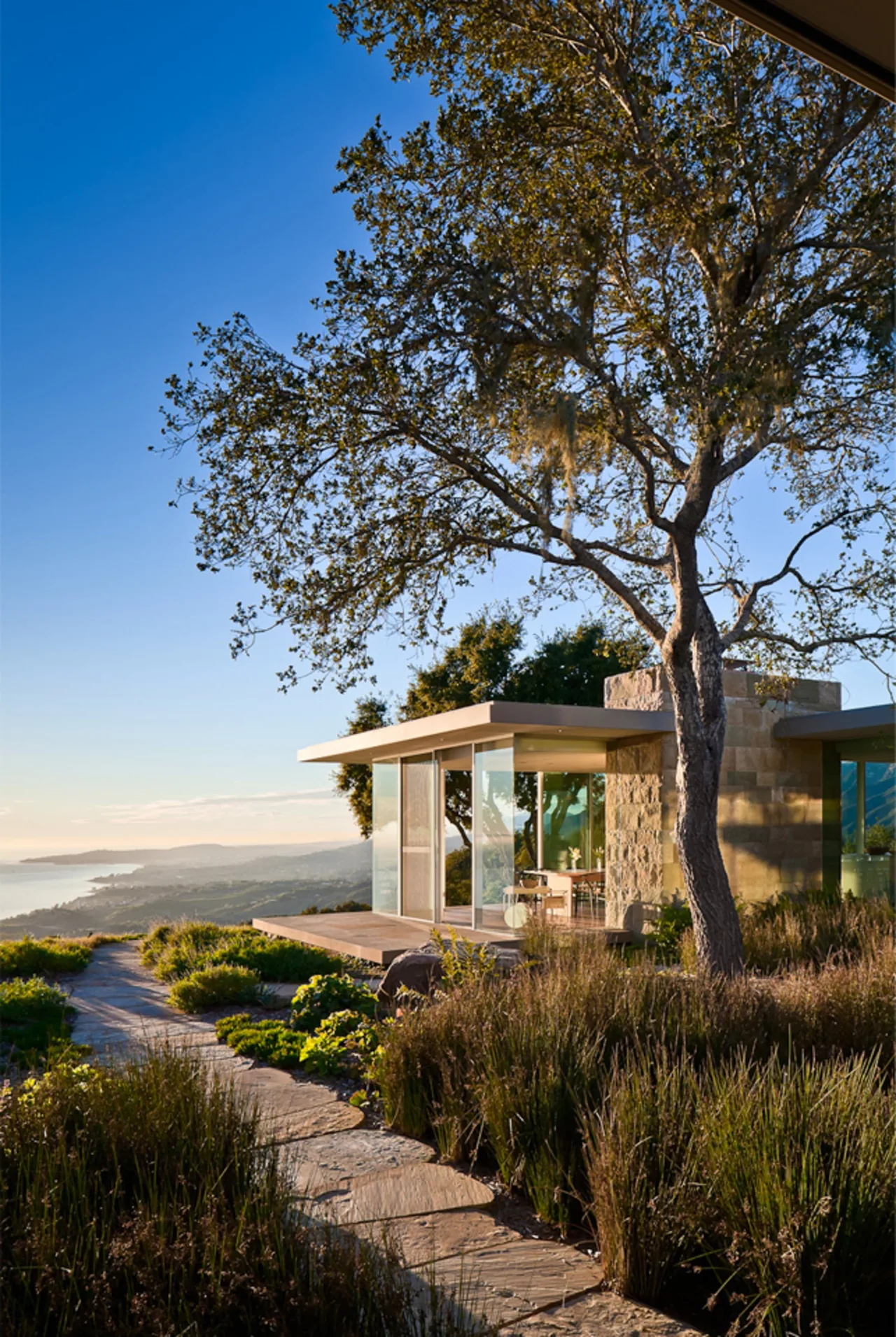 Modern house with flat roof and floor-to-ceiling glass walls overlooking a coastal landscape at sunset.
