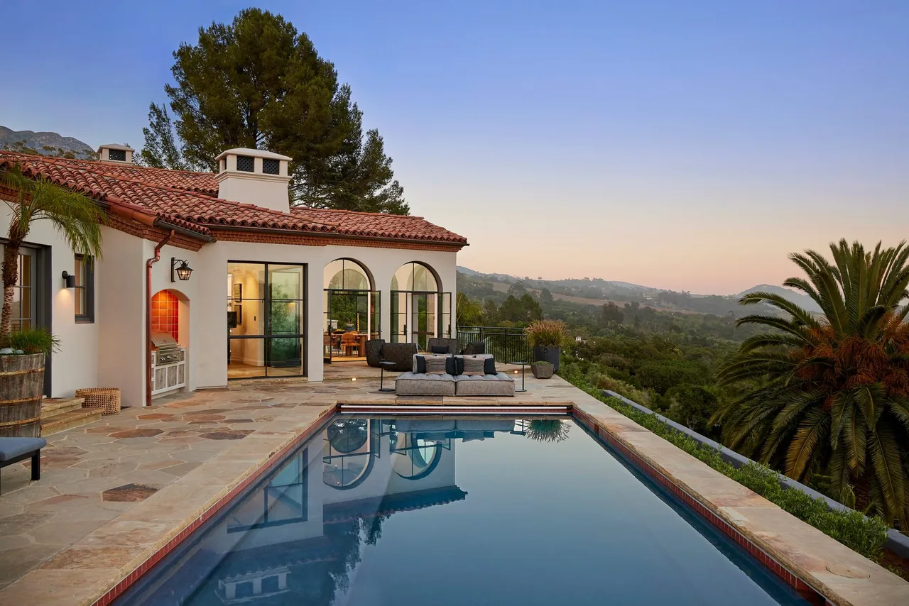 Outdoor patio area with swimming pool, lounge furniture, and a house with terracotta tile roofing at dusk.