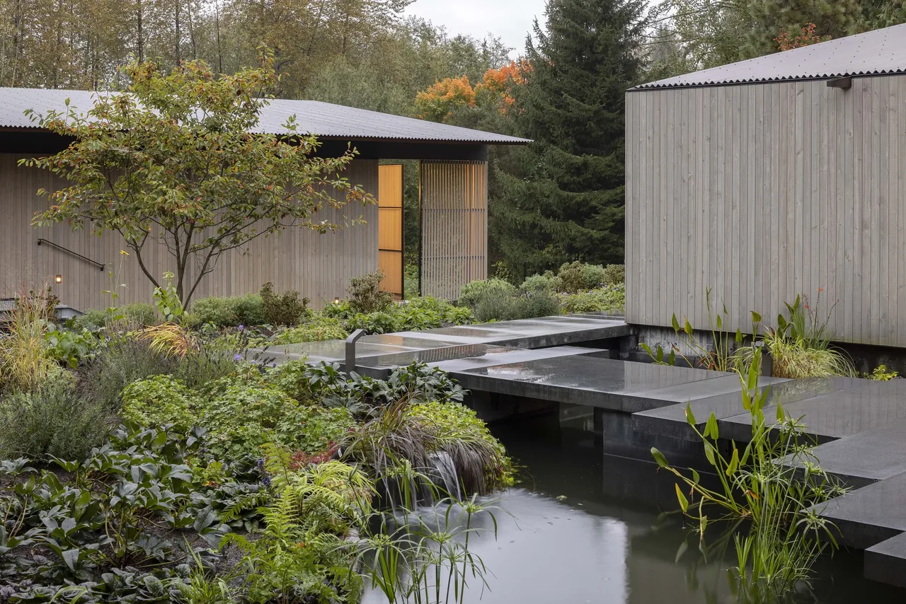 Modern wood-paneled workshop buildings with a concrete stone bridge over a reflective water feature surrounded by greenery.
