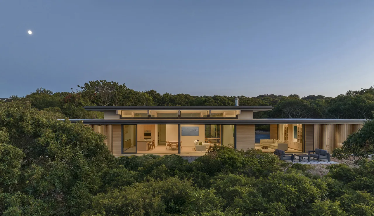 Modern single-story house in wooded area viewed from front at dusk with interior lights on and moon in sky.