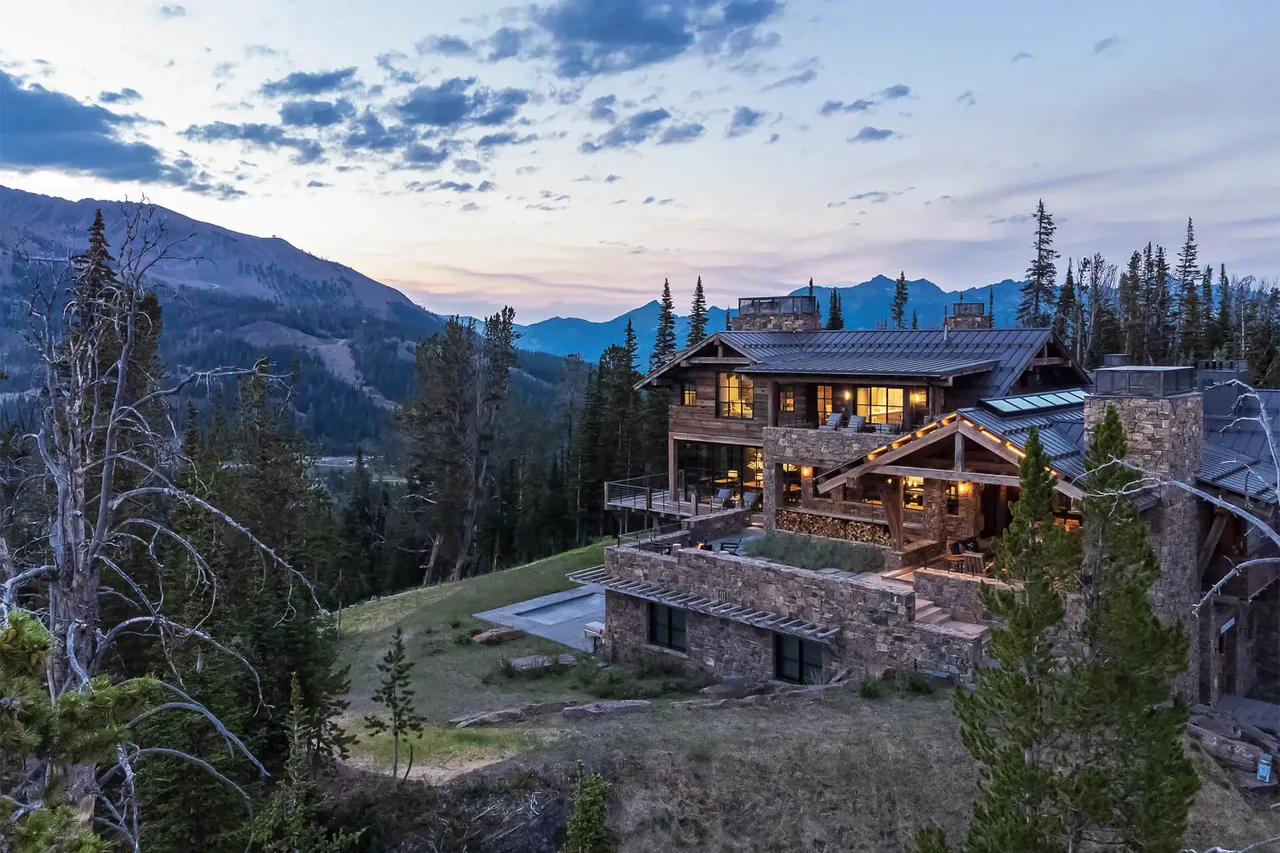 A large rustic mountain house with stone and wood exterior sits on a hillside at sunset surrounded by trees and mountains.