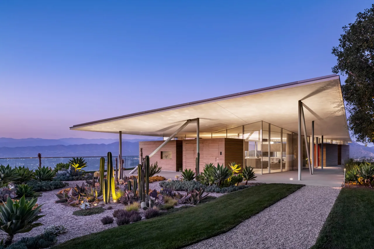 Modern house with a large, angular flat roof and extensive glass walls at twilight surrounded by desert landscaping.