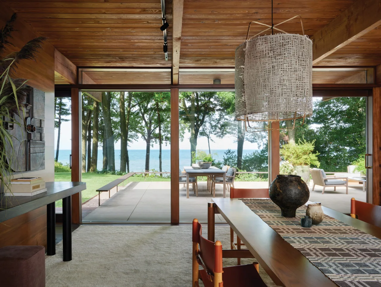 Dining room with large wood table and pendant light, overlooking patio and lake through floor-to-ceiling glass windows.