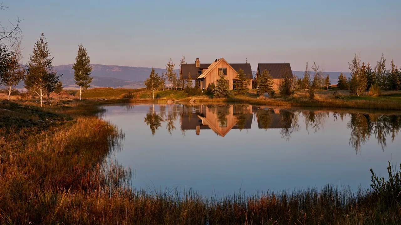 A wooden house reflected in the still water of a pond surrounded by grasses and trees at sunset.