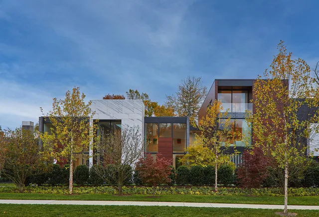 Modern residential building exterior with large windows and autumn trees in front under a blue sky.