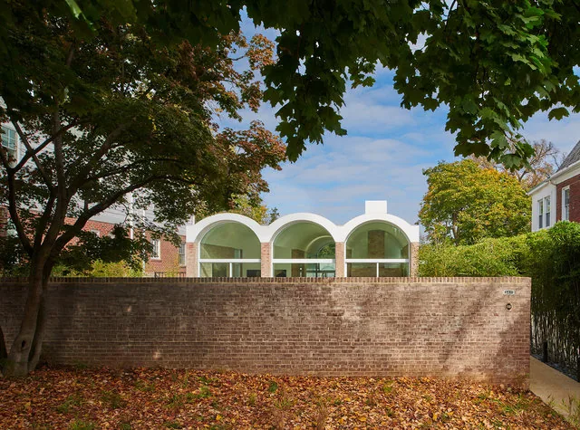 Rear view of a modern house addition with three white vaulted arches above glass walls behind a tall brick wall and autumn trees.