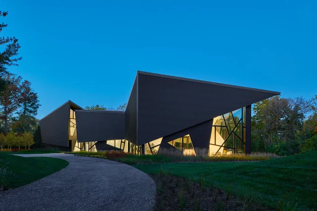 Modern angular house with dark exterior panels and large irregular windows illuminated at dusk